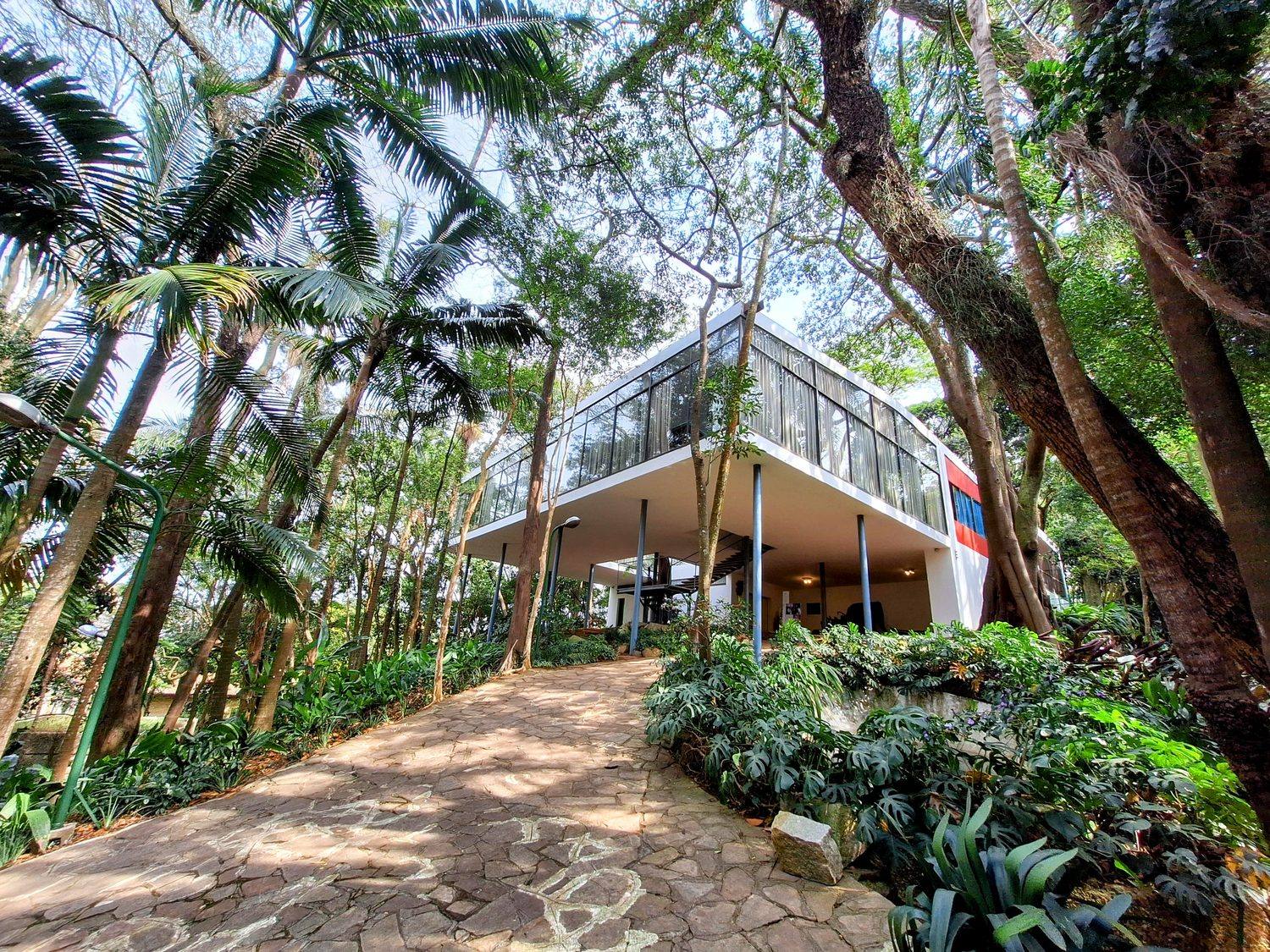 The glass enclosed living level of Casa de Vidro on slender pilotis, seen from the stone path below through the garden.