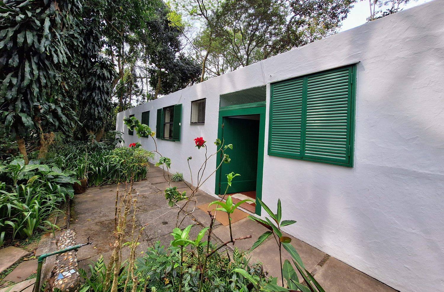 A white-walled garden building of the Casa de Vidro complex with green shutters, set among dense tropical planting.