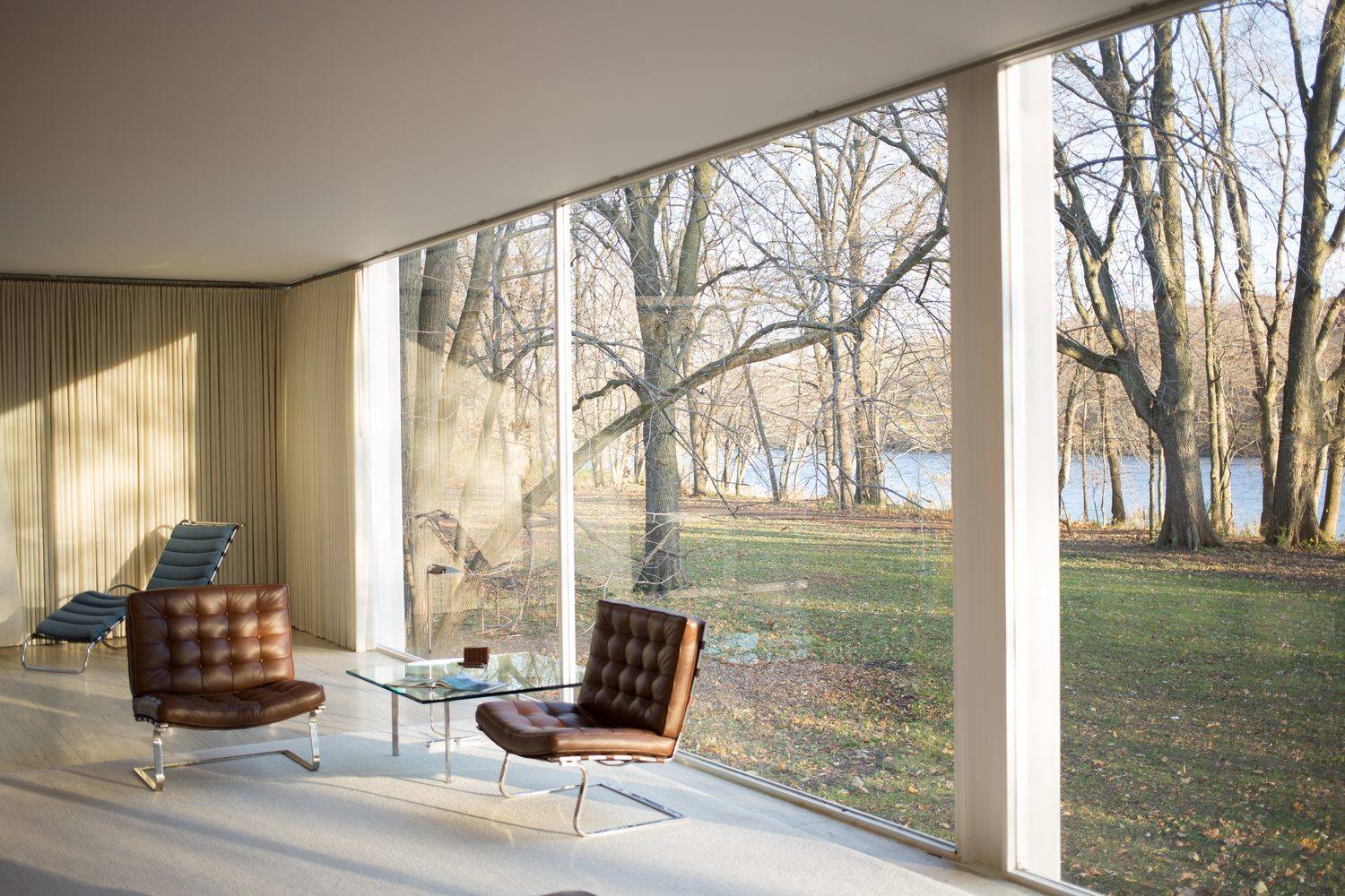 The glass-walled living room of the Farnsworth House with leather lounge chairs, overlooking bare trees and the Fox River.