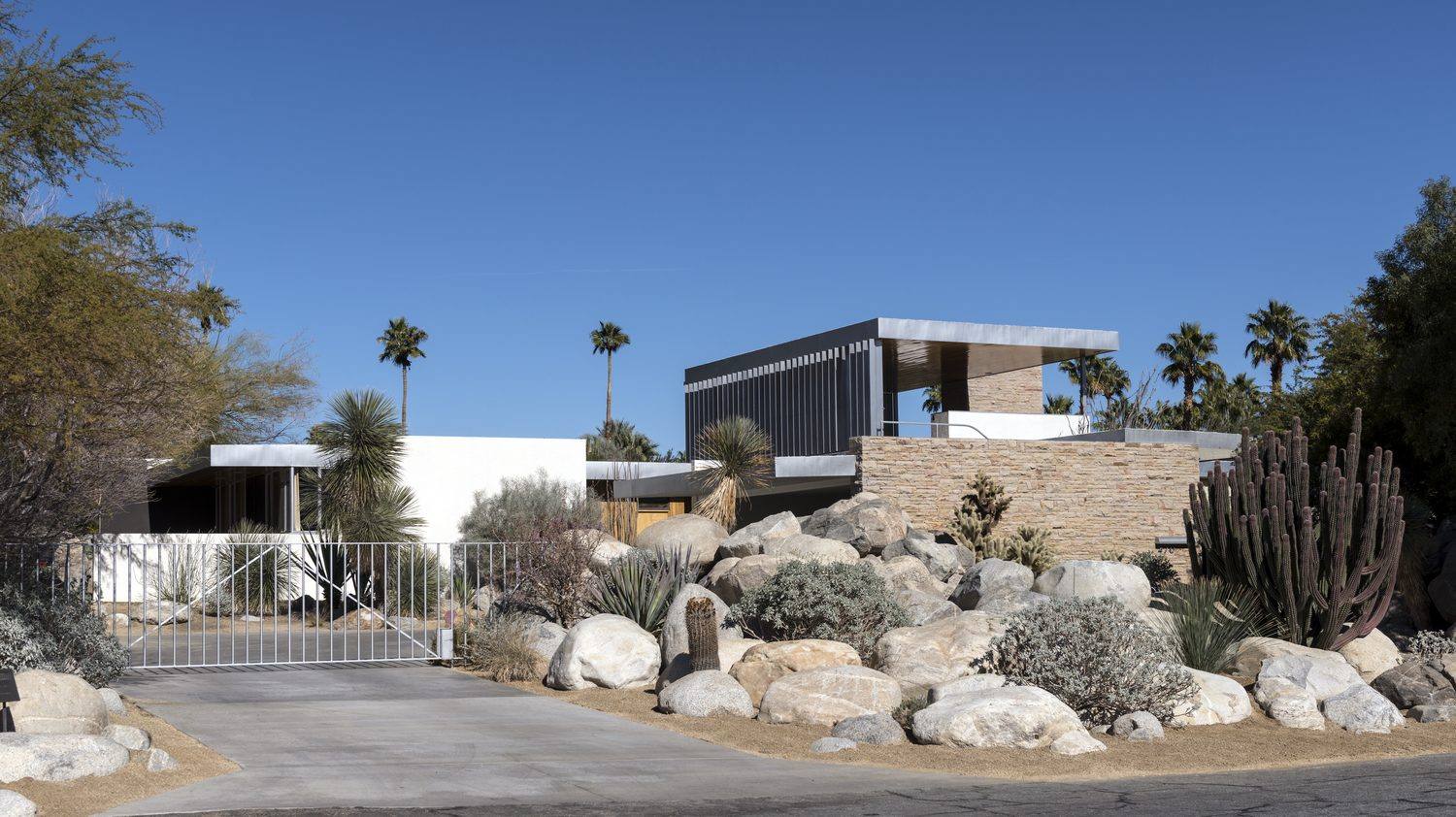 Street view of the Kaufmann Desert House in Palm Springs showing the entry gate, sandstone wall, louvered upper wing and desert rock garden.