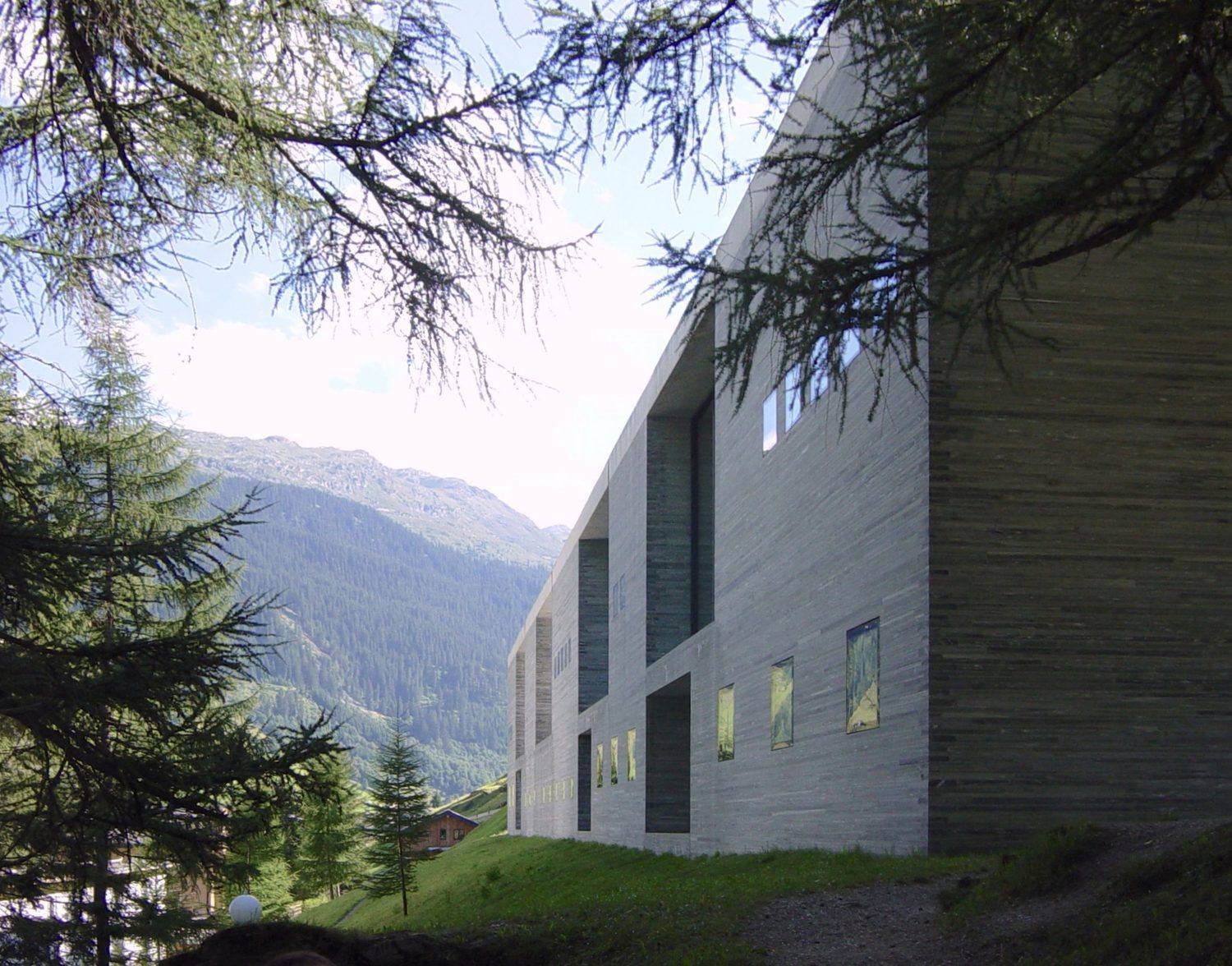 The long quartzite facade of Therme Vals seen through larch branches, with the Alpine slope behind.