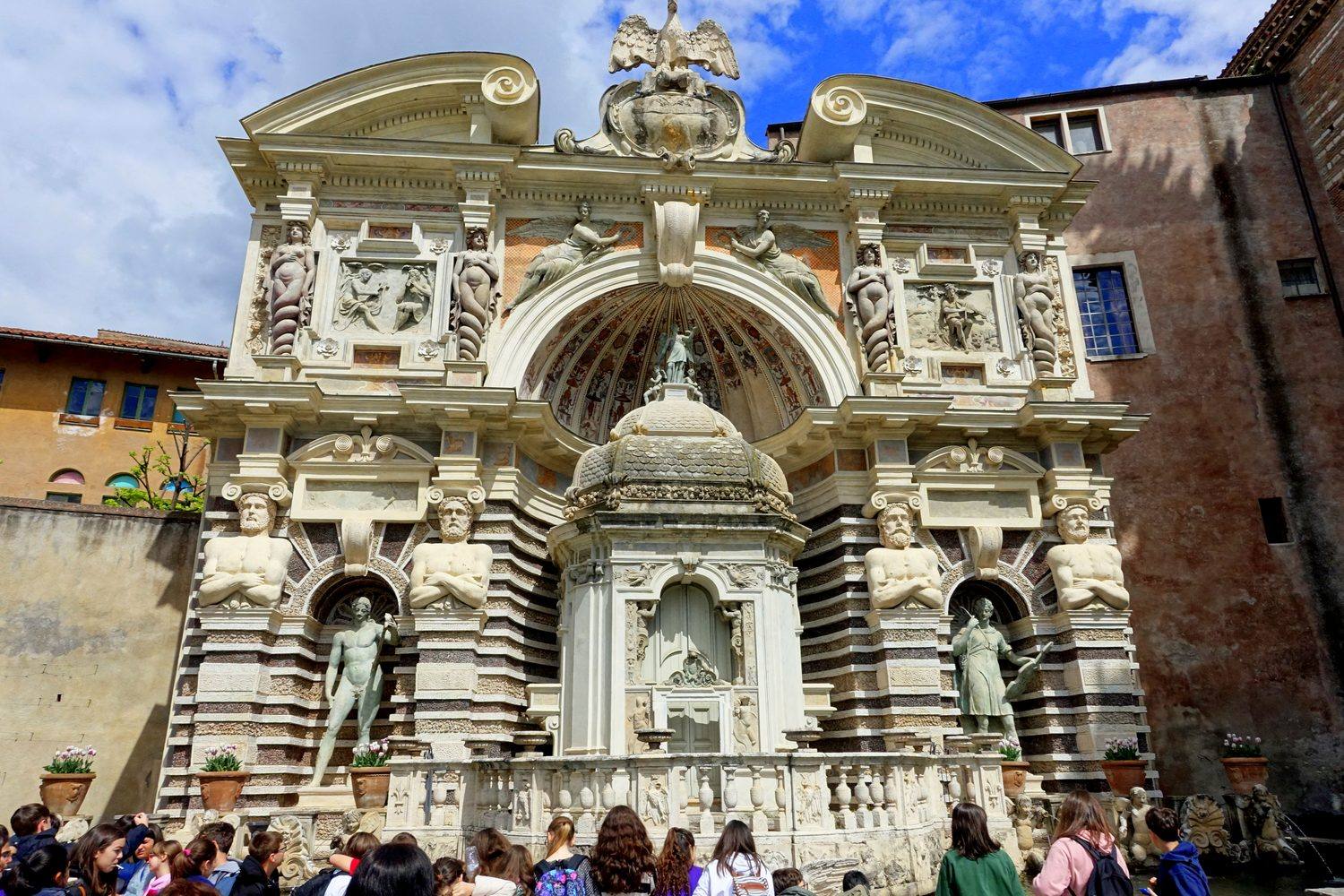 The stone facade of the water organ fountain at Villa d'Este, with sculpted niches and striped masonry, visitors gathered below.